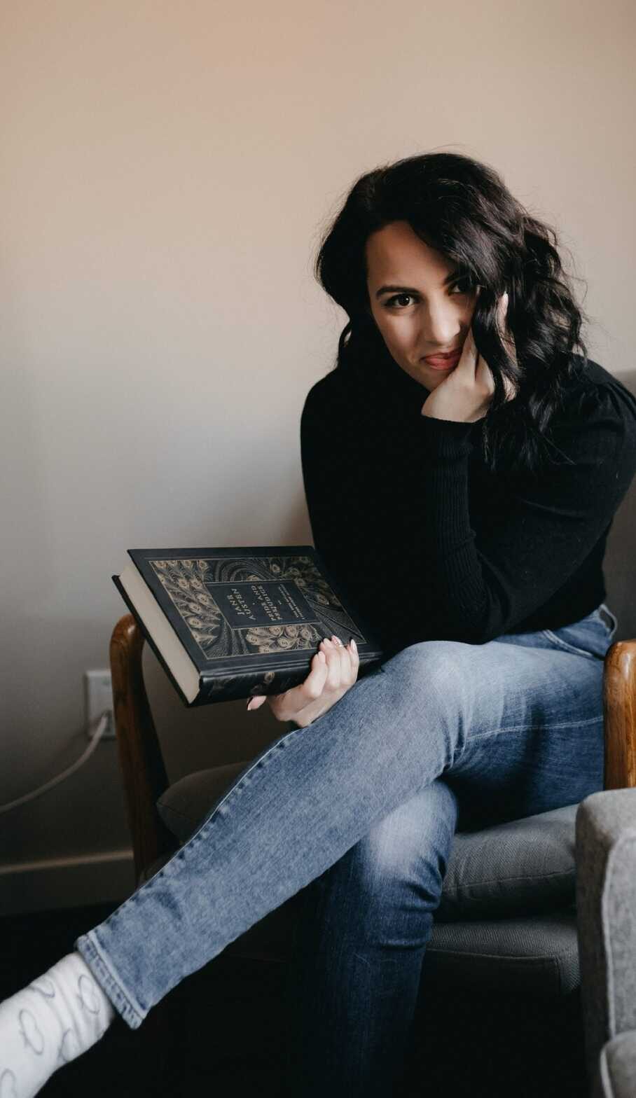 Raegan Milani sits with her legs crossed. She is wearing a black long sleeved shirt and blue jeans. She holds a copy of Jane Austen's Pride and Prejudice.