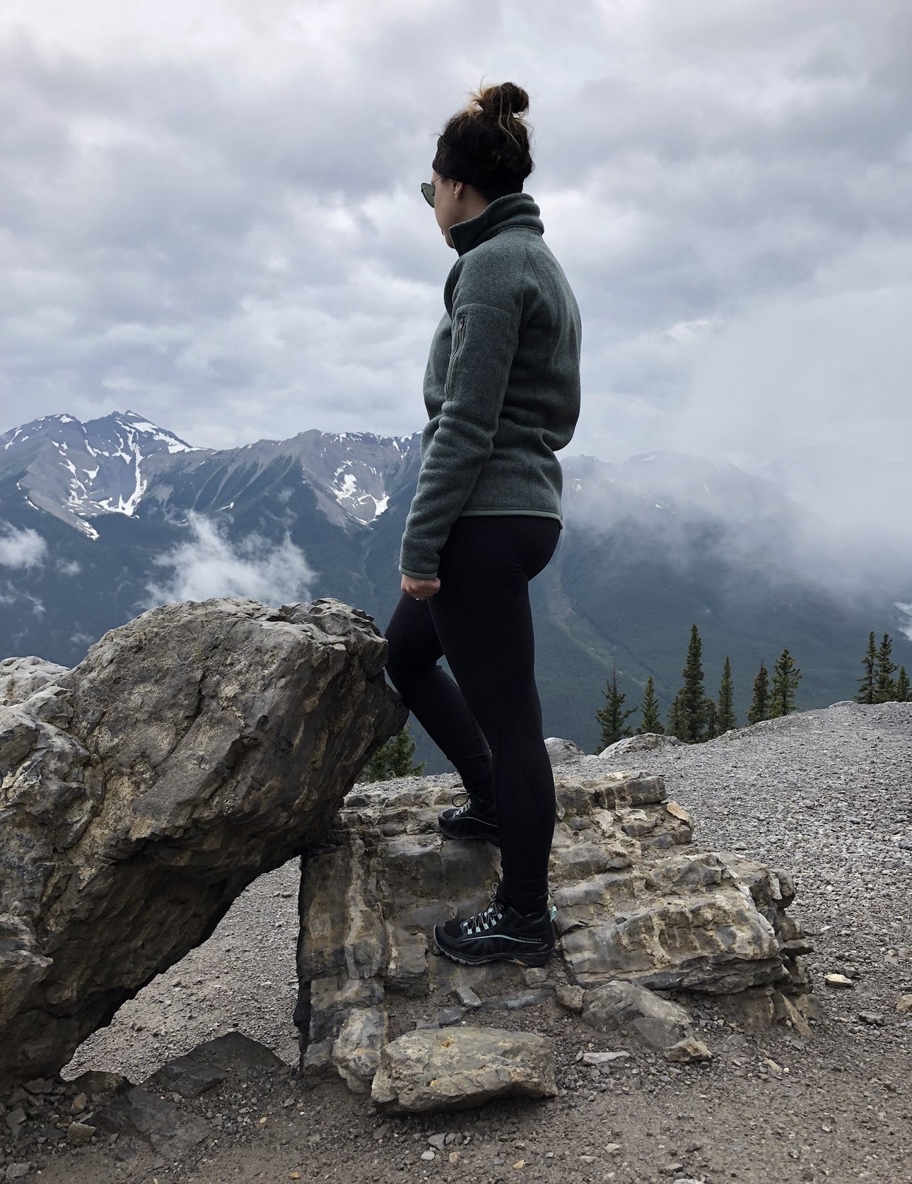 Raegan Milani stands with her back to the camera, facing a mountain. She is wearing black pants, a blueish-green sweater, sunglasses, and hiking shoes.