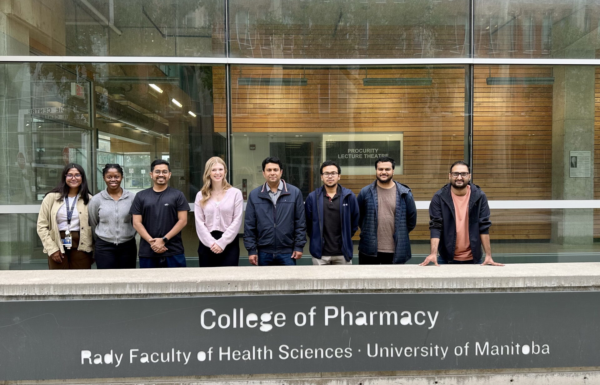 Eight individuals standing behind a sign reading College of Pharmacy, Rady Faculty of Health Sciences, University of Manitoba.