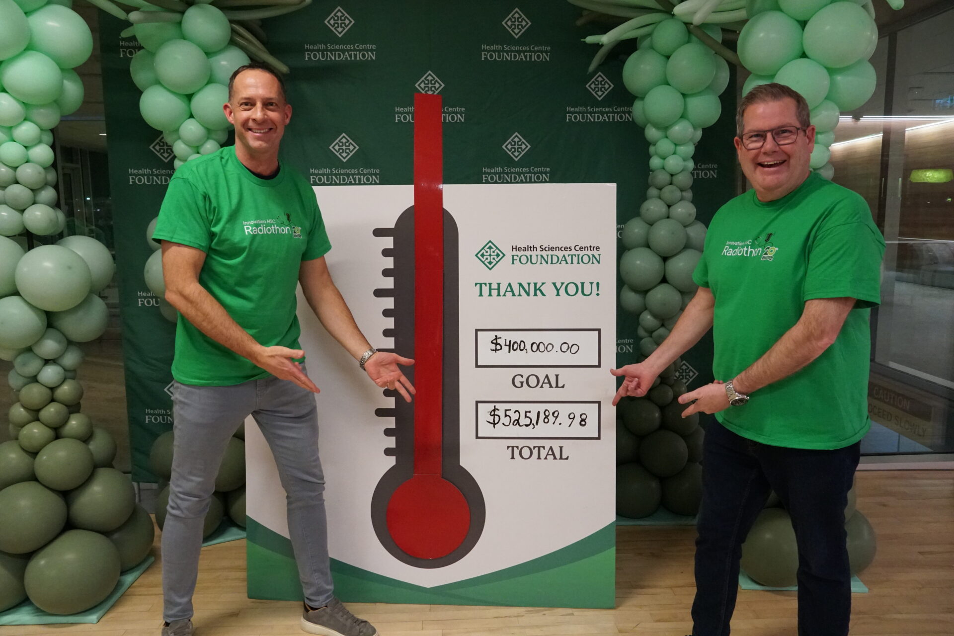 Two smiling men in green shirts stand in front of a sign with a fundraising thermometer showing that the fundraising goal was exceeded and $525,189.98 was raised by the Health Sciences Centre Foundation.